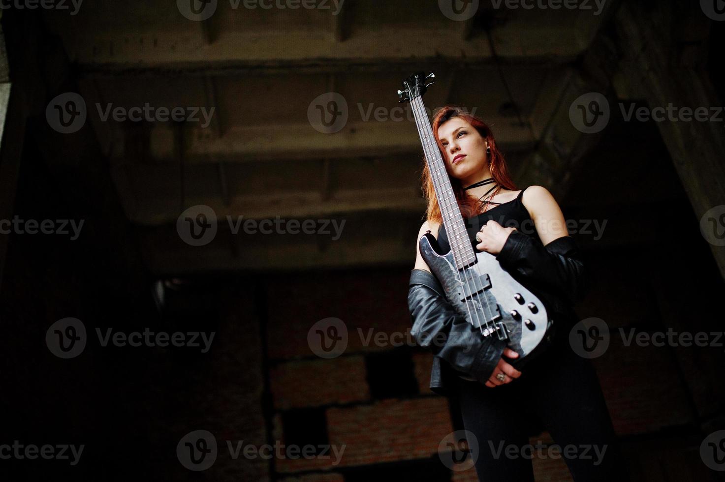 Rothaarige Punkmädchen tragen auf Schwarz mit Bassgitarre an einem verlassenen Ort. Porträt einer gotischen Musikerin. foto