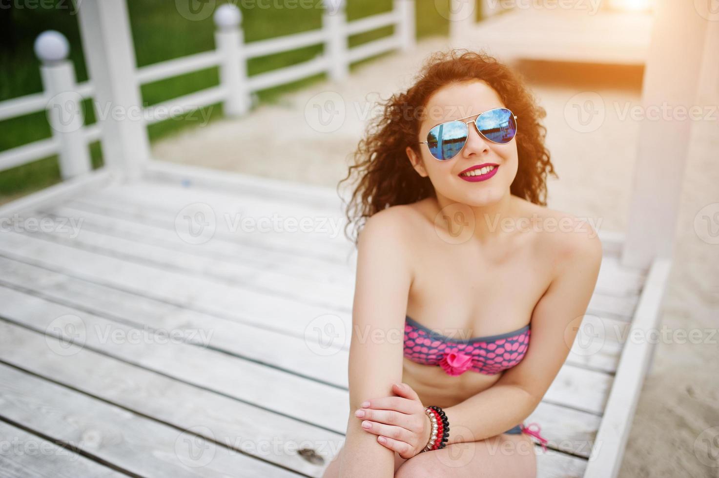 Porträt eines attraktiven Mädchens, das im Bikini mit Sonnenbrille posiert, während es auf einer weißen Holzterrasse steht. foto