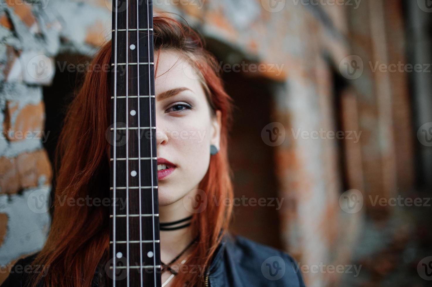 Rothaarige Punkmädchen tragen auf Schwarz mit Bassgitarre an einem verlassenen Ort. Porträt einer gotischen Musikerin. nahaufnahme gesicht der schwärze person mit gitarrenriff. foto