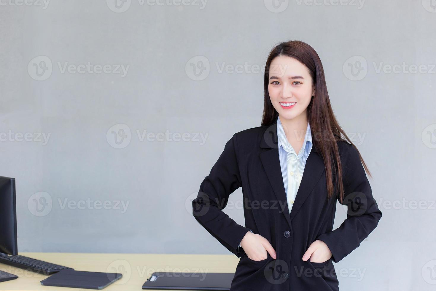 professionelles Geschäft arbeitende Asiatin steht und steckt ihre Hand in die Tasche eines schwarzen Anzugs, während sie im Büro als Hintergrund glücklich lächelt. foto