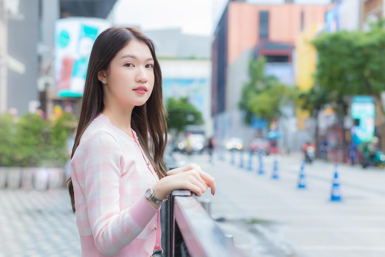 schöne asiatische frau in einem rosa-weiß gestreiften mantel steht im städtischen freien mit der stadt und der straße als hintergrund. foto