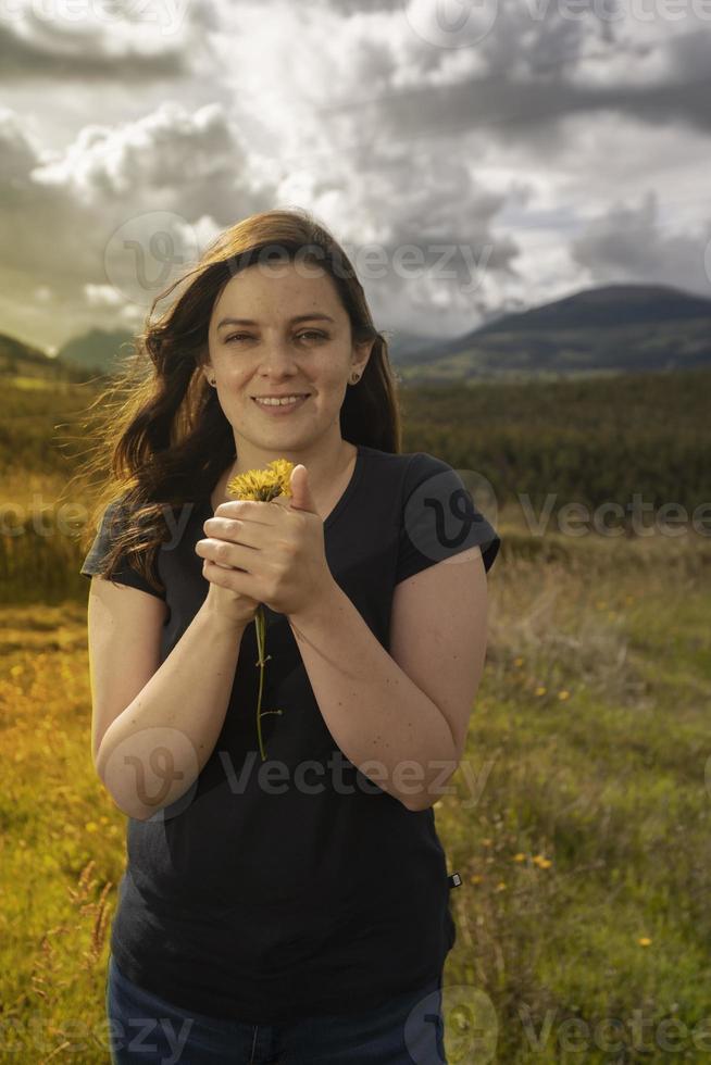 Schöne glückliche hispanische Frau, die der Kamera in der Mitte des Feldes gegenübersteht und gelbe Blumen riecht, die sie in ihren Händen hält foto