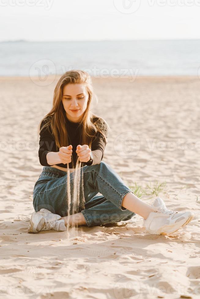 Schöne Frau in voller Länge, die abends bei Sonnenuntergang am Strand sitzt und Sand gießt. hübsches Weib foto