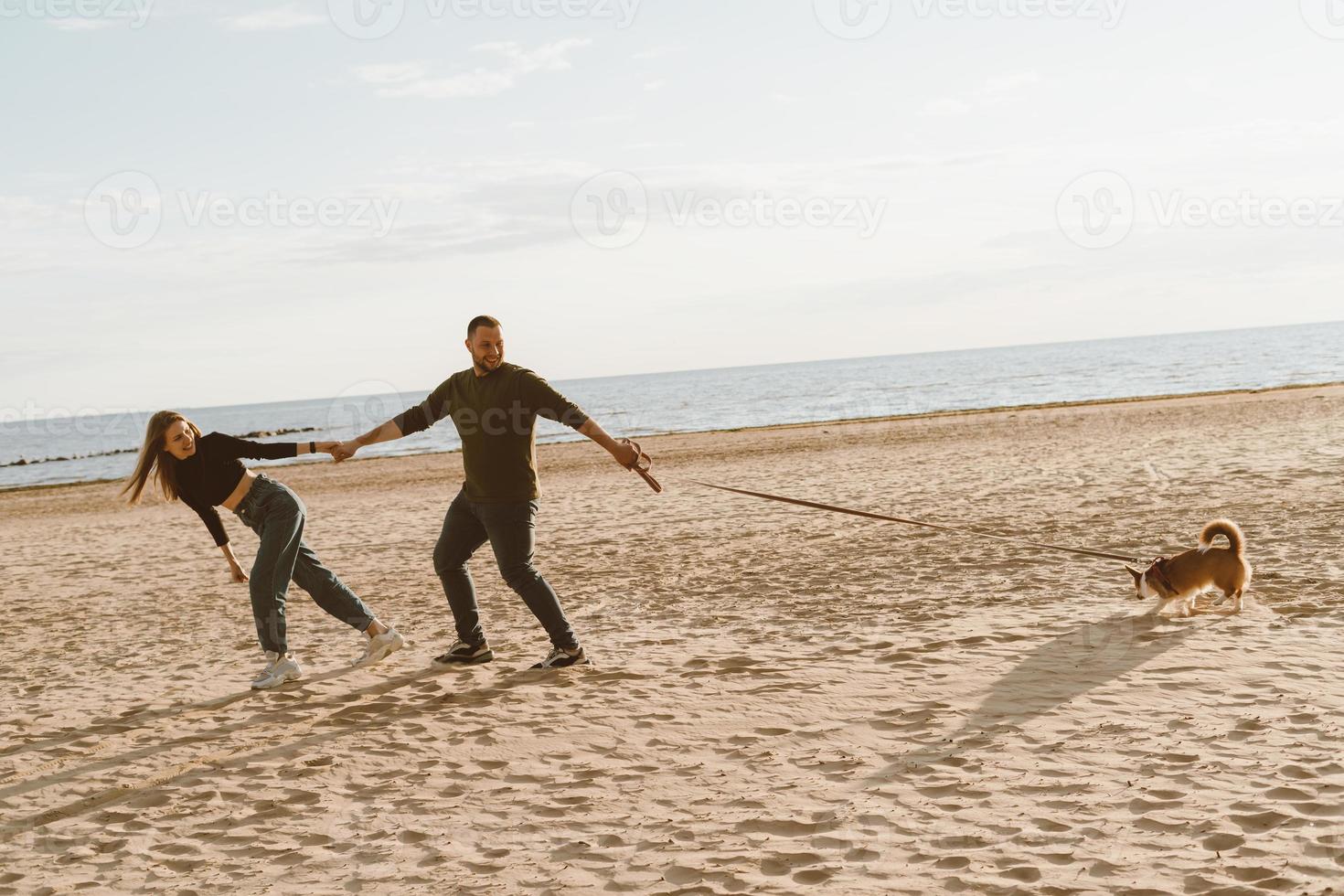 junges glückliches paar und hund, die am strand spazieren gehen. gutaussehender Mann, der schöne Frau hält foto