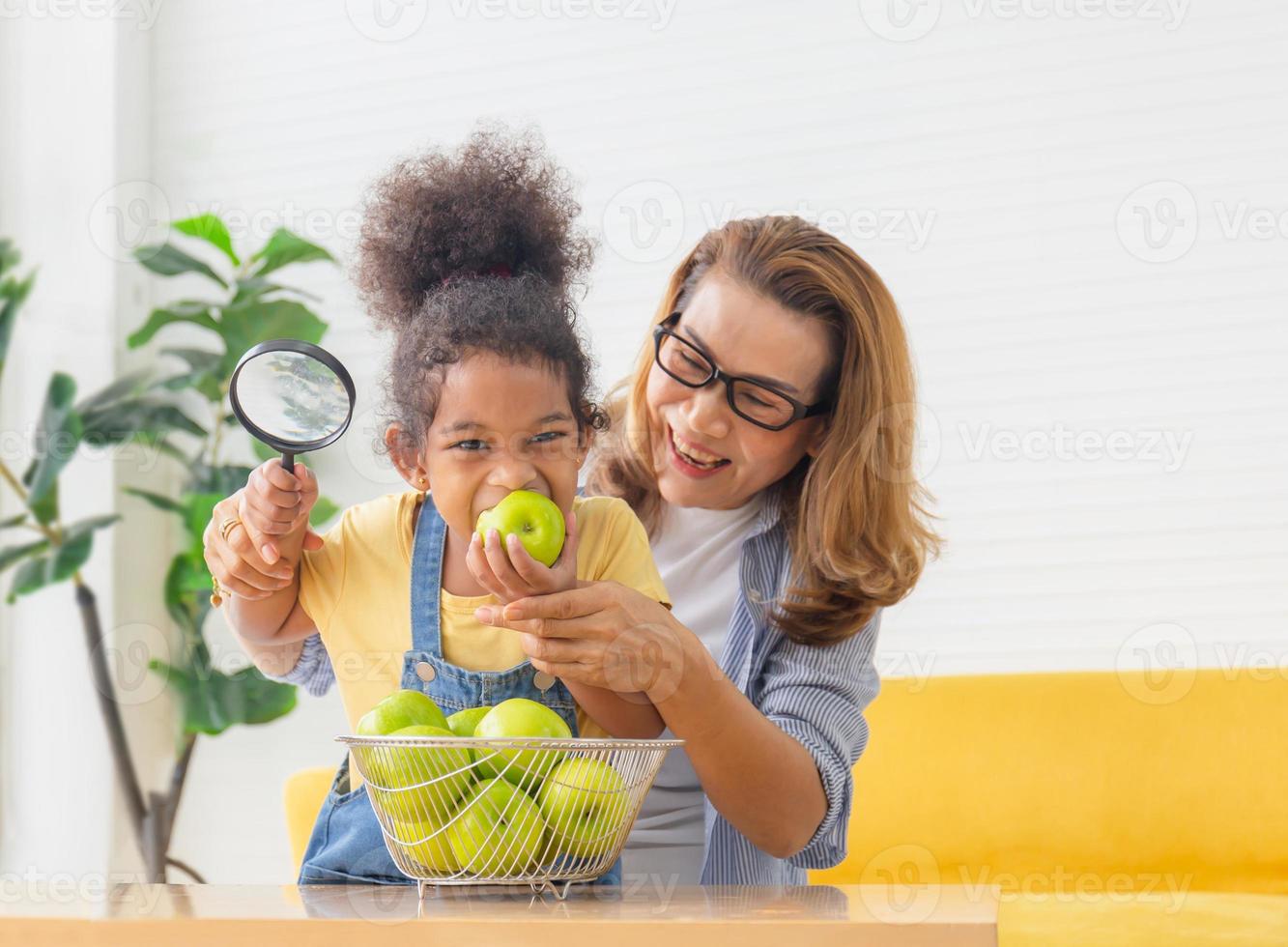 Fröhliche Tochter und Mutter mit Apfelfrucht in der Küche, kleines Mädchen und Großmutter mit Lupe spielen fröhlich im Wohnzimmer foto