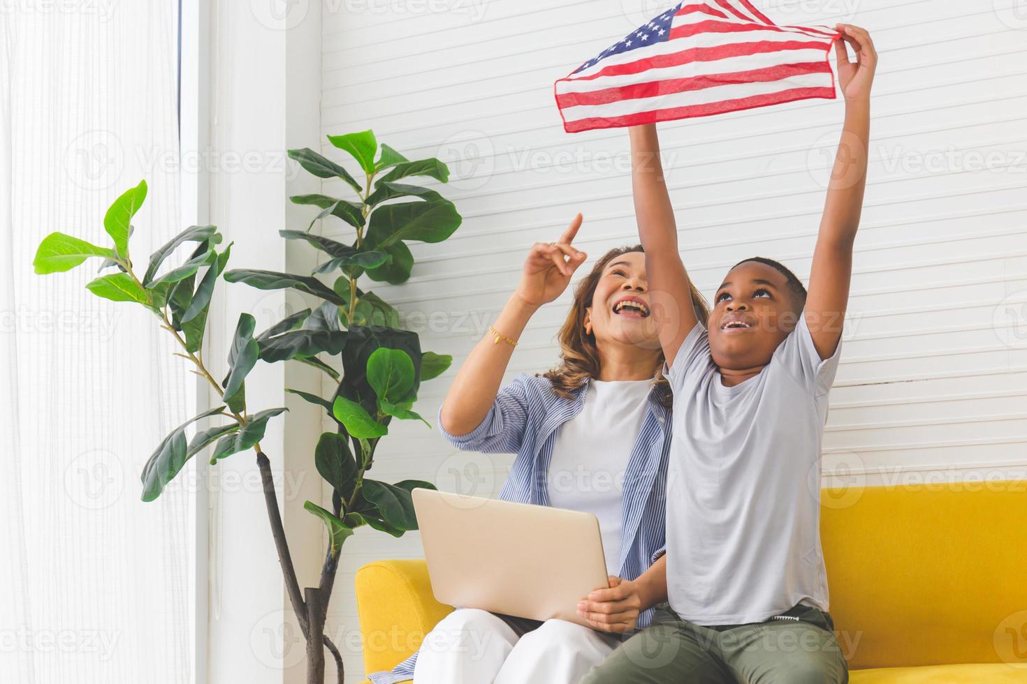 Fröhliche reife Frau mit Laptop aussehender kleiner Junge mit USA-Flagge, Großmutter und Enkelkindern, die fröhlich im Wohnzimmer spielen foto