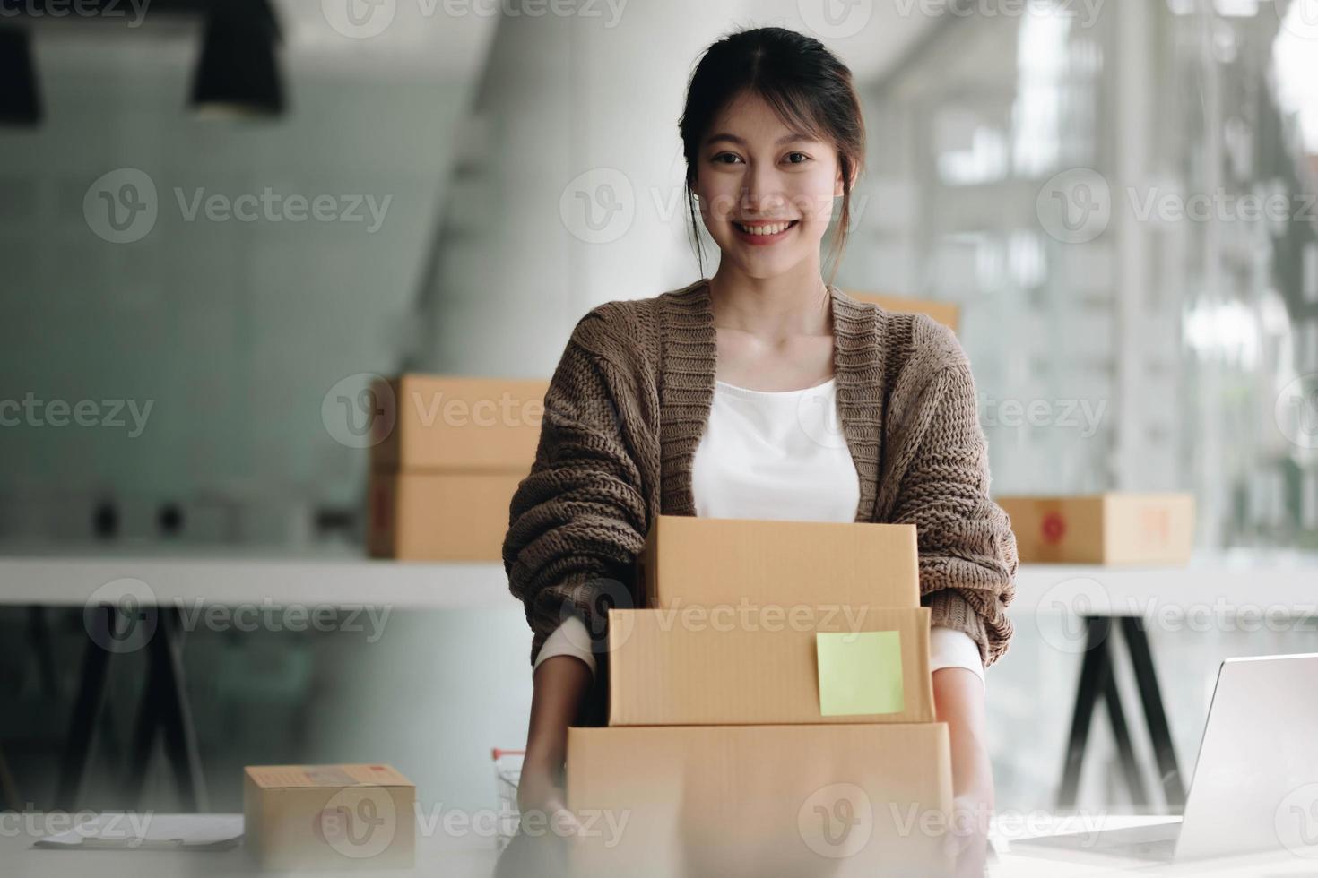 glückliche junge asiatische frau start kleinunternehmen freiberuflich tätig hält paketbox und computer laptop. lieferkonzept für online-marketing-verpackungsboxen foto