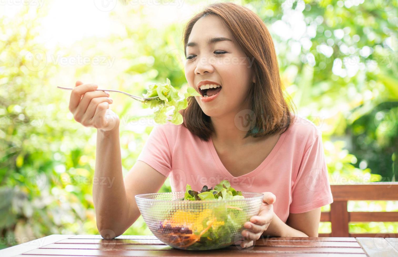 die glückliche schöne asiatische gesunde frau mittleren alters sitzt auf dem balkon des hauses neben dem garten und isst einen gesunden grünen salat. konzept der gesundheitsversorgung und nahrhaften lebensmittel foto