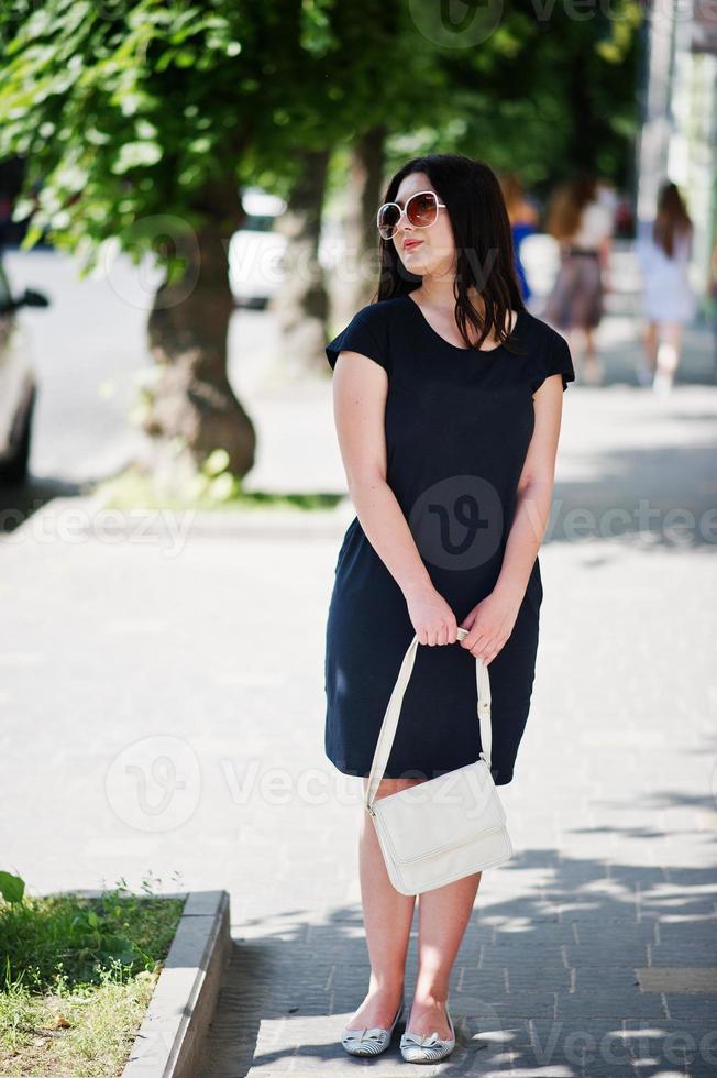 brünettes mädchen im schwarzen kleid auf sonnenbrille mit handtasche zur hand, die auf der straße der stadt posiert. foto