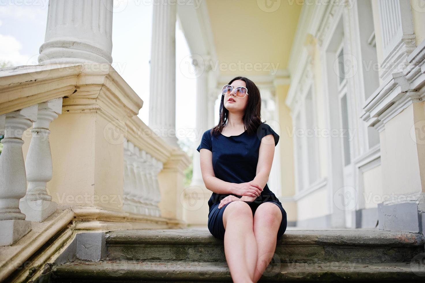 Brünettes Mädchen im schwarzen Kleid, Sonnenbrille, die auf der Treppe eines alten Vintage-Hauses sitzt und auf der Straße der Stadt posiert. foto