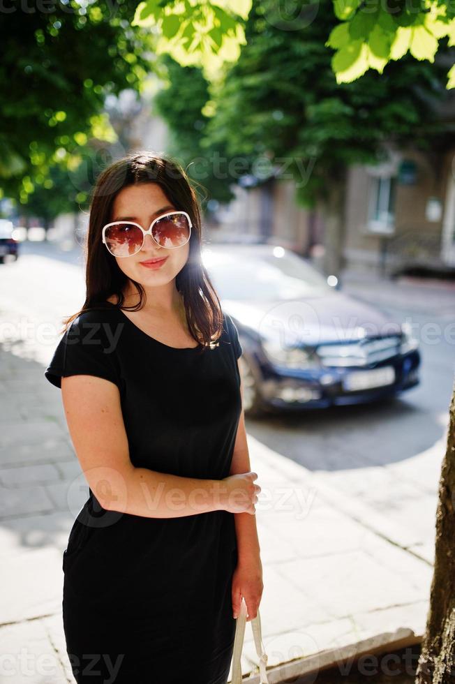 brünettes Geschäftsmädchen im schwarzen Kleid auf Sonnenbrille mit Handtasche zur Hand posiert auf der Straße der Stadt, Hintergrundauto auf der Straße. foto