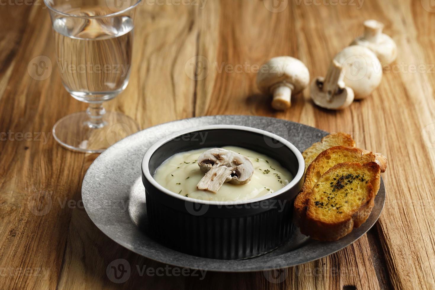 Champignoncremesuppe mit knusprigem Knoblauchbrotbaguette, serviert auf schwarzer Keramikschale oder Auflaufförmchen über rustikalem Tisch foto