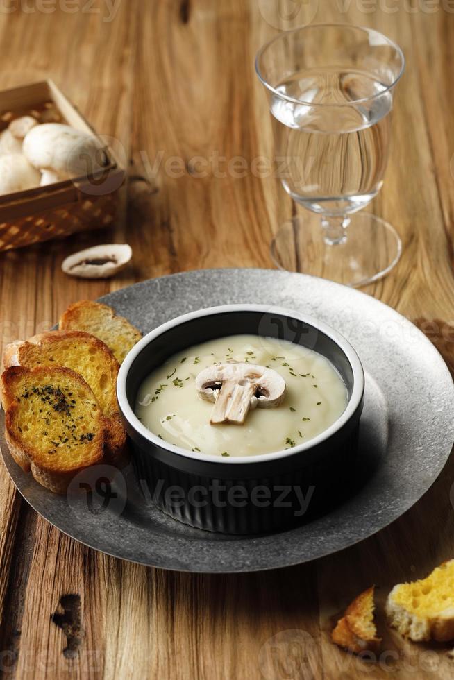 Pilzcremesuppe mit knusprigem Knoblauchbrotbaguette, serviert auf schwarzer Keramikschale oder Ramequin, auf rustikalem braunem Holztisch. foto