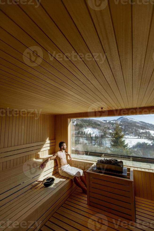 junger mann, der sich in der sauna entspannt und den winterwald durch das fenster beobachtet foto