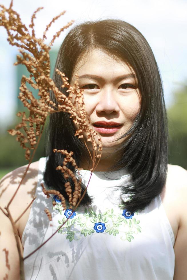 schöne asiatische frau im weißen kleid lächelnd und hand, die trockene zweige im naturpark hält. thailändisches mädchen oder chinesisches mädchen genießen im urlaub mit sonnenlicht im garten foto