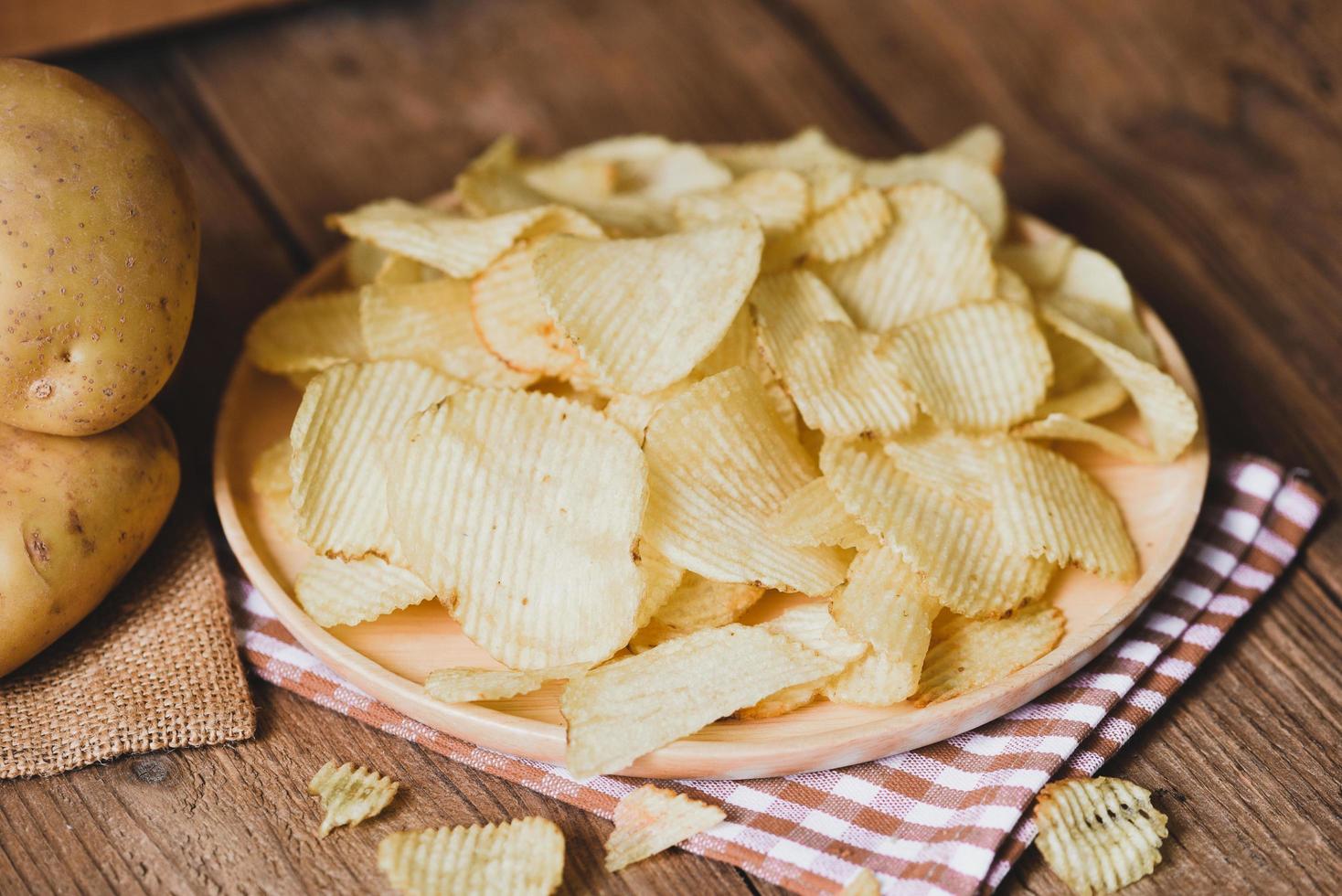 Kartoffelchips-Snack auf weißem Teller, knusprige Kartoffelchips auf dem Küchentisch und frische rohe Kartoffeln foto