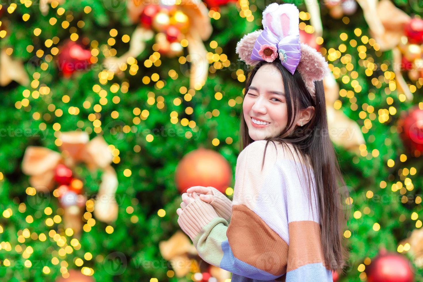 ein asiatisches mädchen in einem bunten pullover steht vor dem weihnachtsbaum. mit bokeh als hintergrund im thema des feierns des neuen jahres foto