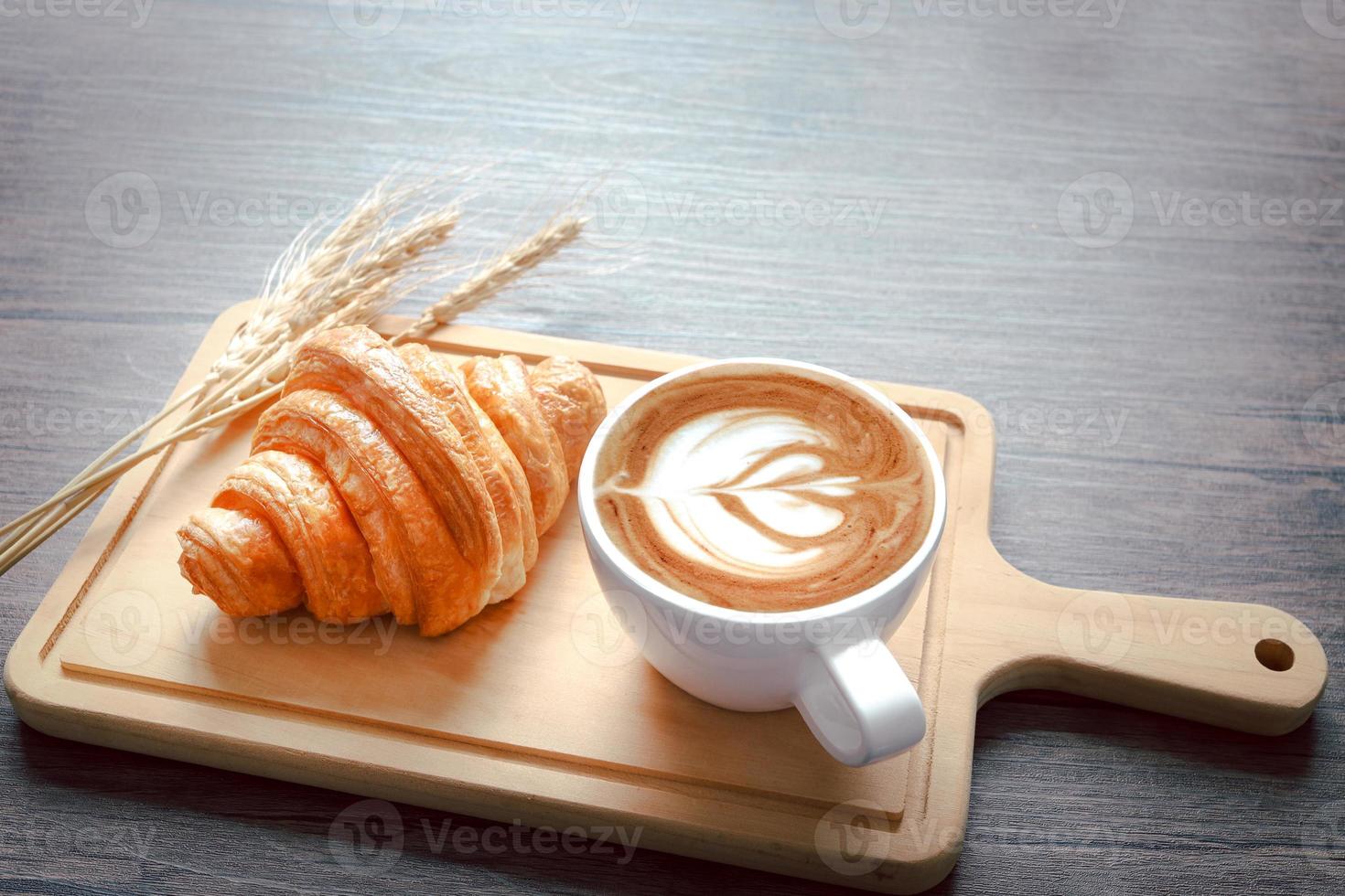 frisch gebackenes leckeres Croissant mit Ährchen und Tasse schönen Morgenkaffee auf Holzschneidebrett, Essen auf dem Frühstückstisch foto