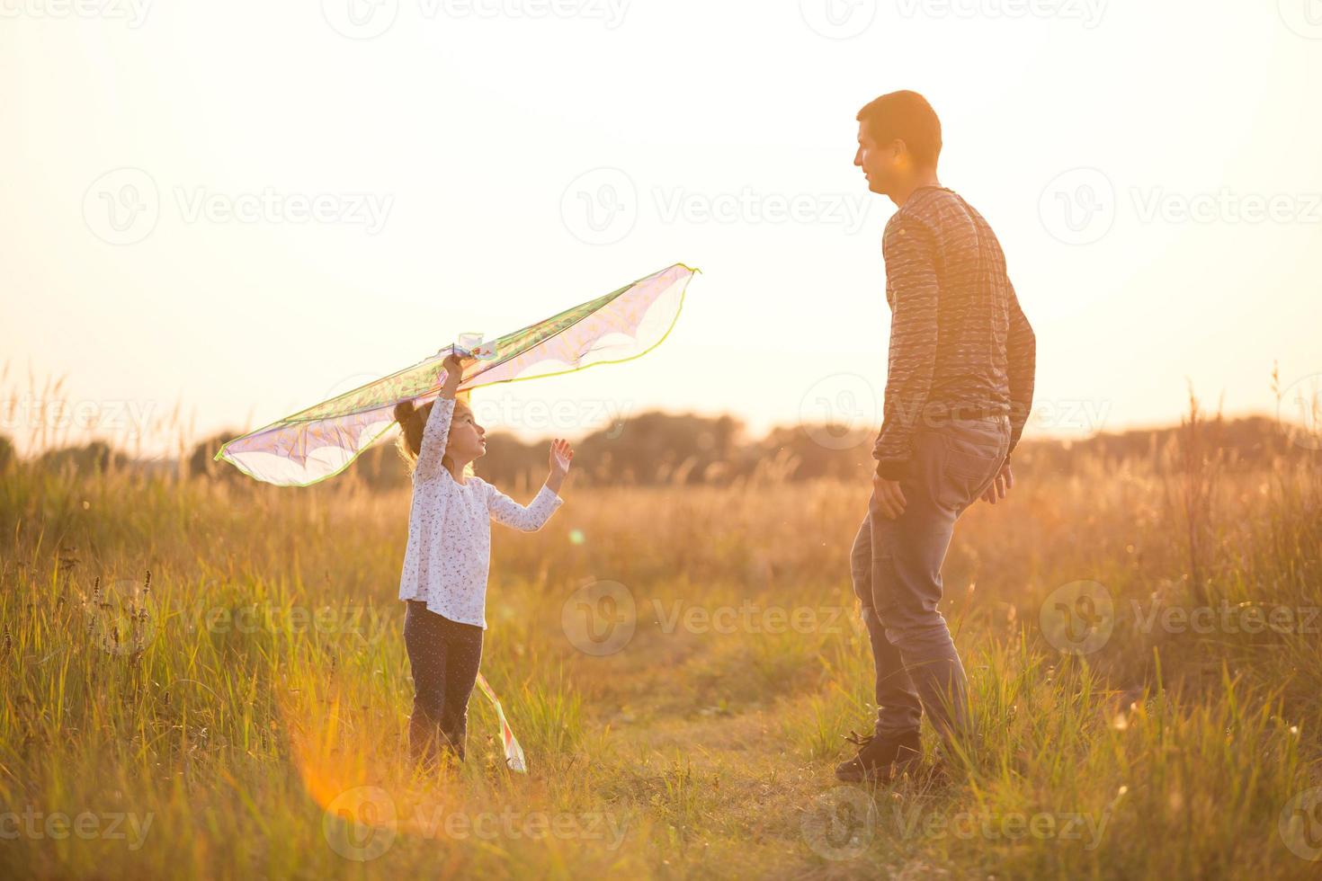 Papa hilft seiner Tochter, im Sommer bei Sonnenuntergang einen Drachen auf einem Feld zu steigen. Familienunterhaltung im Freien, Vatertag, Kindertag. ländliche Gebiete, Unterstützung, gegenseitige Hilfe. orangefarbenes Licht der Sonne foto