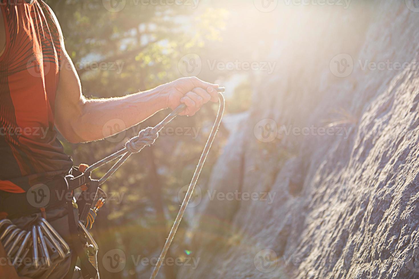 Die Hand eines männlichen Kletterers hält ein Seil mit einem Sicherheitsknoten von acht. Ein Mann in Kletterausrüstung auf dem Hintergrund von Felsen im Licht der Sonne. Karabiner, Hosenträger, Expressschlingen, Gürtel, Geschirr. Exemplar foto