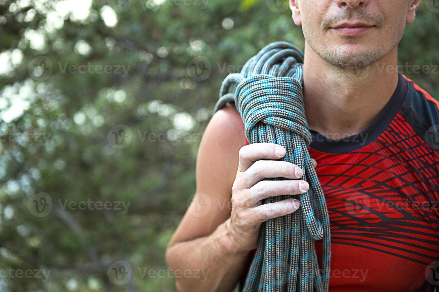 Ein männlicher Kletterer von kaukasischem Aussehen mit einem aufgerollten Seil auf der Schulter. Hand in weiße Magnesia. rotes Sporttrikot, Extremsport, Klettern. Platz für Text foto