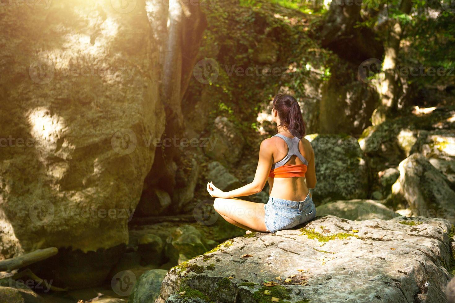 Eine Frau sitzt in einem Lotusfeld auf einem großen Felsen zwischen den Felsen im Freien und meditiert, genießt die Einheit mit der Natur, lauscht der Stille und den Geräuschen des Waldes. Ökologie foto