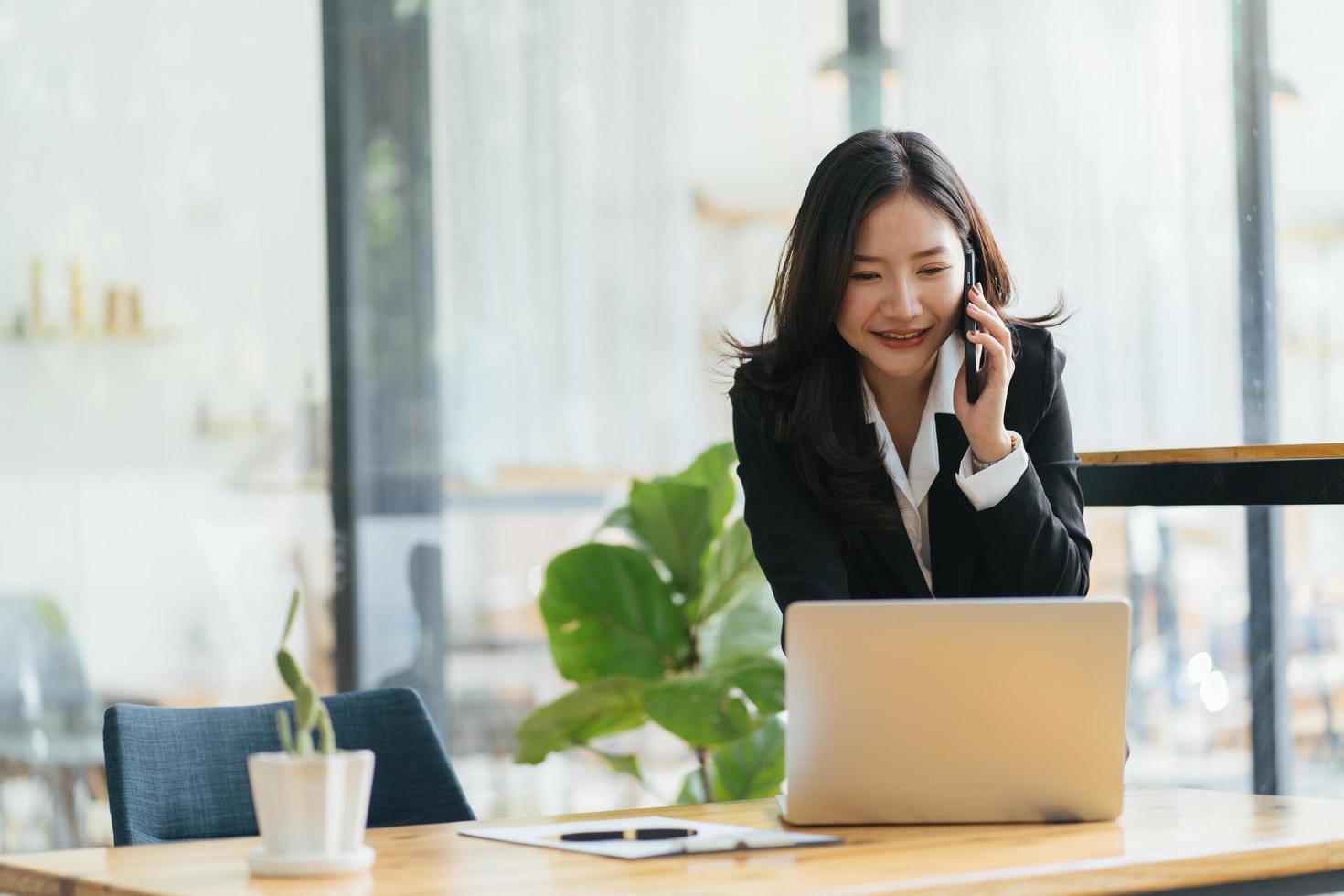 glücklich lächelnde asiatische Geschäftsfrau, die im Büro am Laptop arbeitet und ein Smartphone verwendet. Geschäftsfrau sitzt an ihrem Arbeitsplatz foto