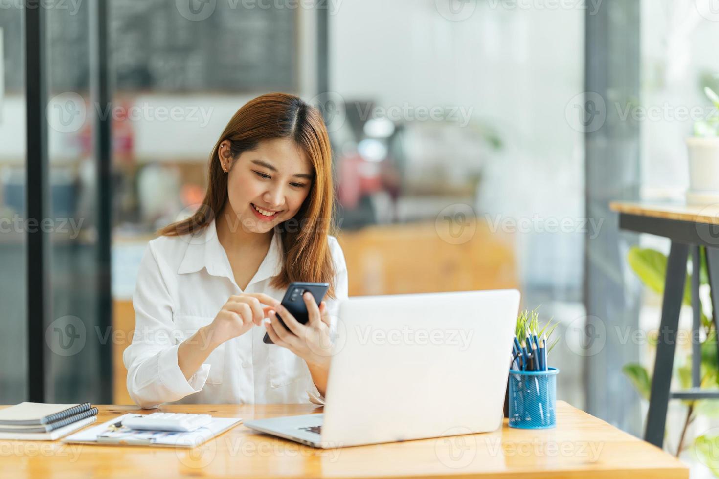 asiatische frau in lässiger kleidung ist glücklich und fröhlich, während sie mit ihrem smartphone kommuniziert und in einem café arbeitet. foto