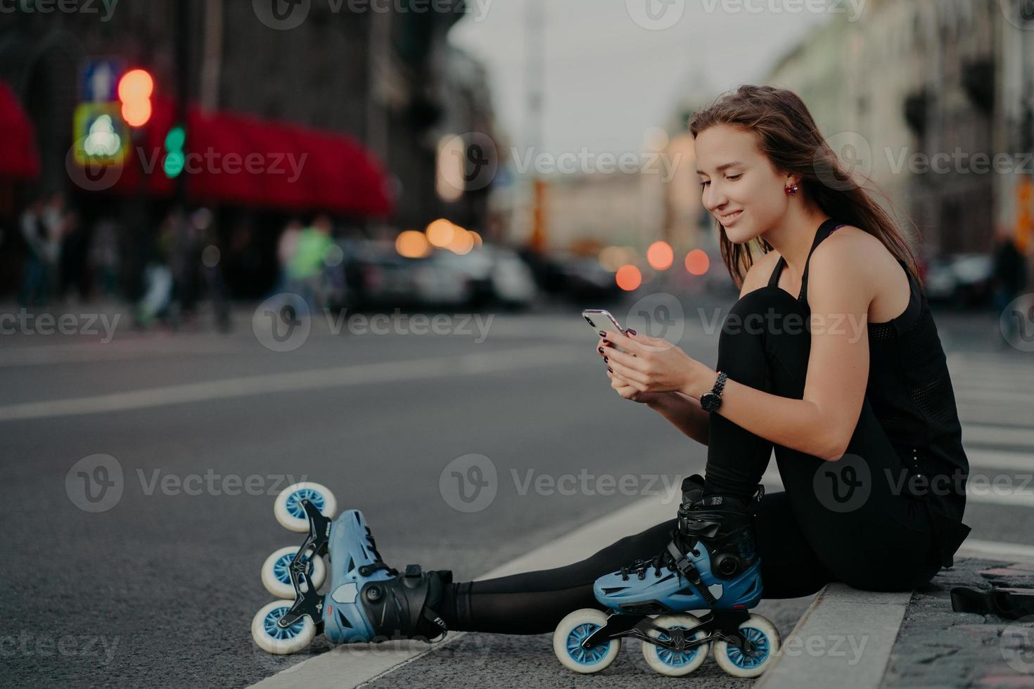 Zufriedene sportliche Frau trägt Sportkleidung, Rollerblades, sitzt auf der Straße, überprüft den Newsfeed per Smartphone, macht eine Pause, nachdem Inline-Skaten vor verschwommenem Stadthintergrund posiert und sich für einen gesunden Lebensstil einsetzt foto