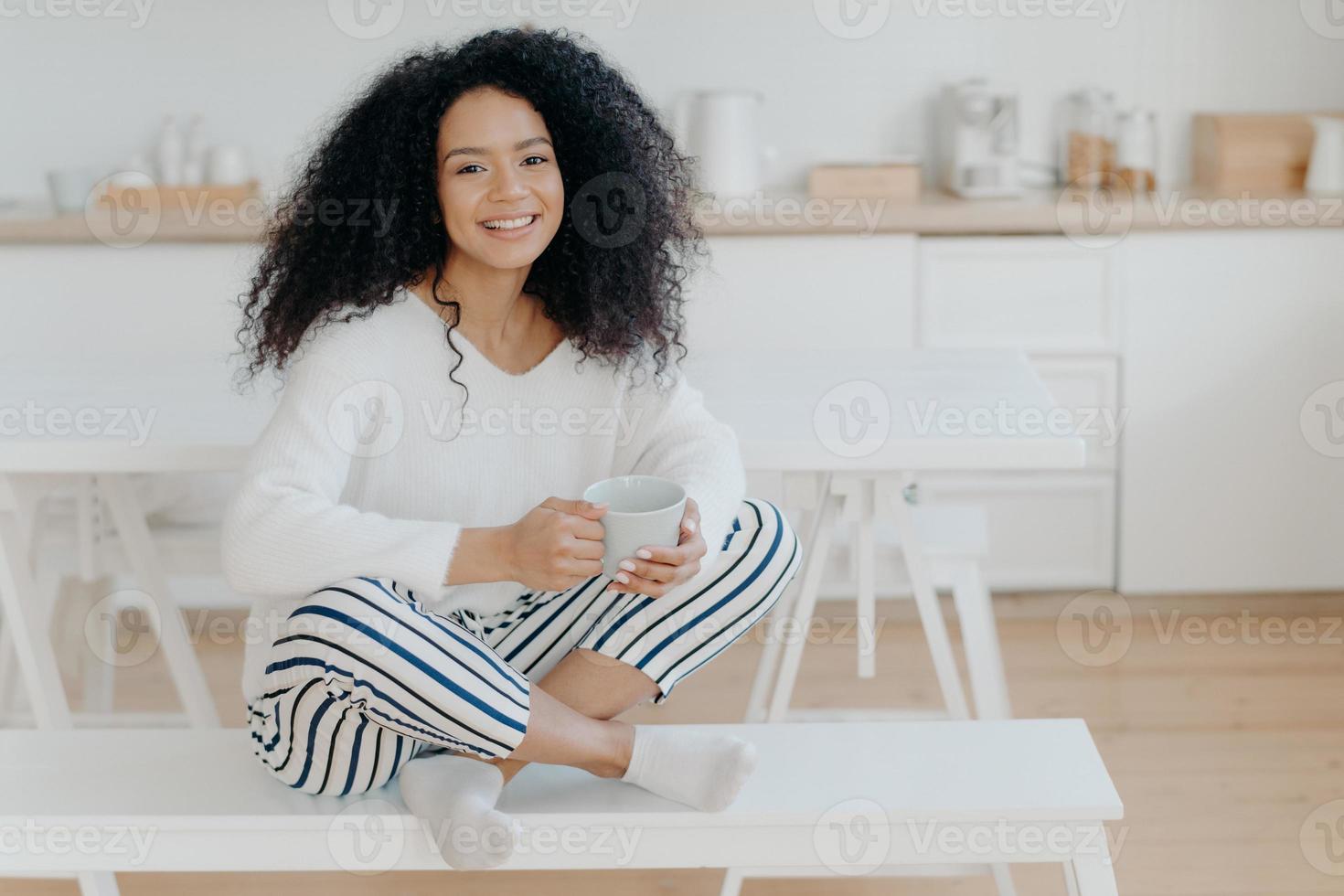 fröhliches morgenkonzept. Foto einer fröhlichen, lockigen Afroamerikanerin sitzt in Lotus-Pose auf einer weißen Bank, nippt an einem leckeren aromatischen Getränk, fühlt sich entspannt, posiert vor dem Kücheninterieur und lächelt breit