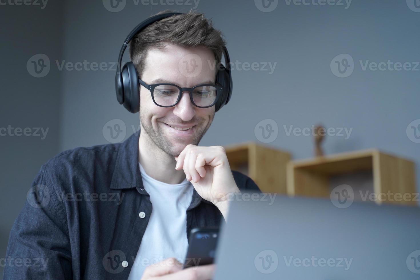 glücklicher Heimbüroangestellter mit Kopfhörern und Brille, der aufrichtig lächelt, wenn er auf den Telefonbildschirm schaut foto