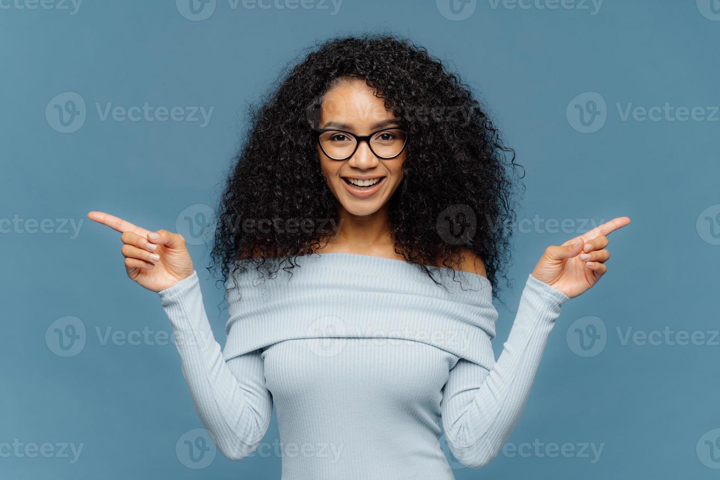 glückliche frau mit afrofrisur, zeigt mit beiden zeigefingern zur seite, trägt einen lässigen pullover, zögert zwischen zwei objekten, isoliert auf blauem hintergrund. weiblich zeigt links und rechts innen an foto