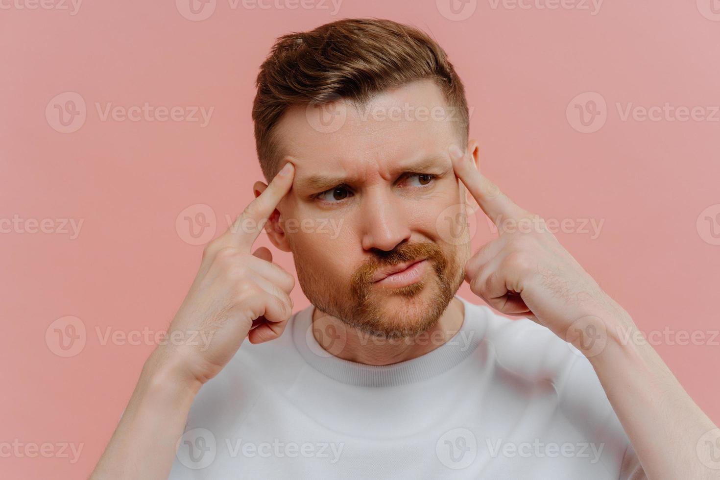 Nahaufnahme eines ernsthaften bärtigen erwachsenen Mannes hält die Finger an den Schläfen konzentriert beiseite und versucht, sich an etwas im Kopf zu erinnern, das lässig isoliert über rosafarbenem Hintergrund gekleidet ist. männliches Model denkt tief foto