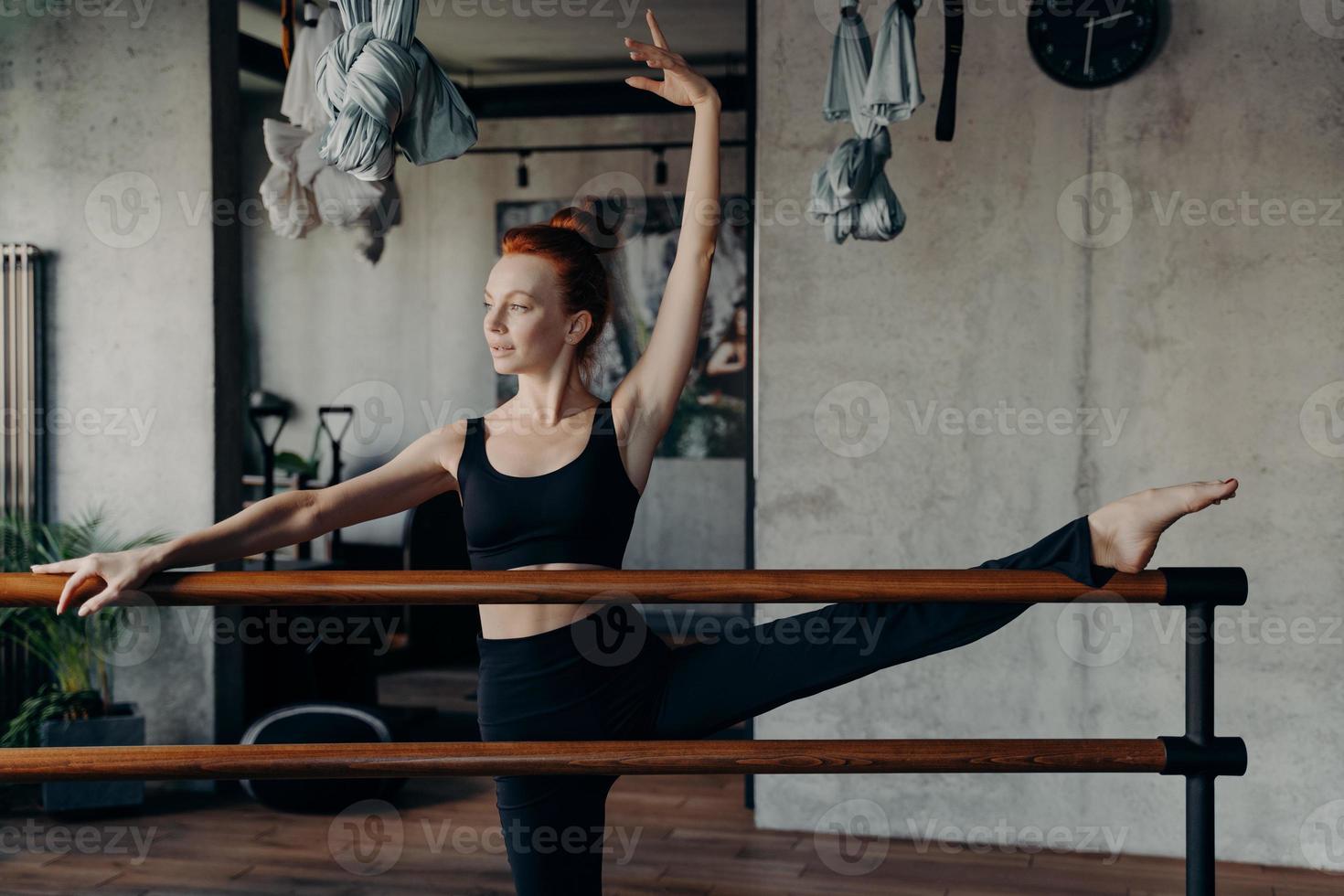 junge athletische rothaarige ballerina, die sich im fitnessstudio das bein auf der ballettstange dehnt foto