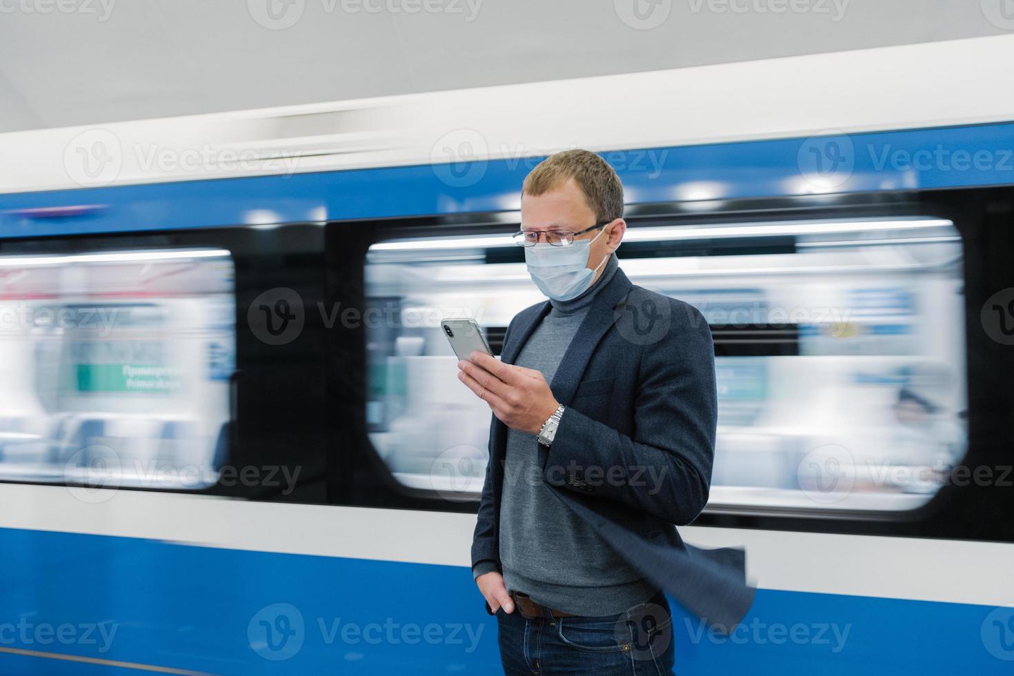 Foto eines Mannes mit Brille und medizinischer Schutzmaske, benutzt ein modernes Telefon, fährt mit öffentlichen Verkehrsmitteln, stellt sich auf dem Bahnsteig gegen den fahrenden Zug, informiert sich online über das neue Covid-19-Virus