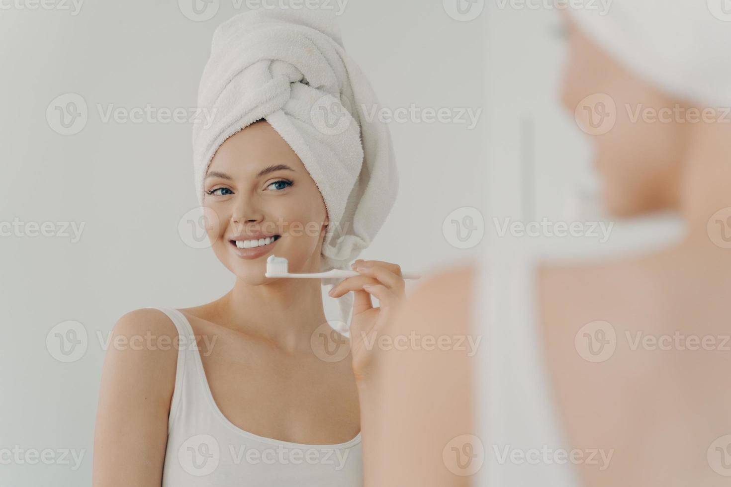 Schöne junge Frau mit gesundem, perfektem Lächeln, die Zähne putzt und in den Spiegel schaut foto