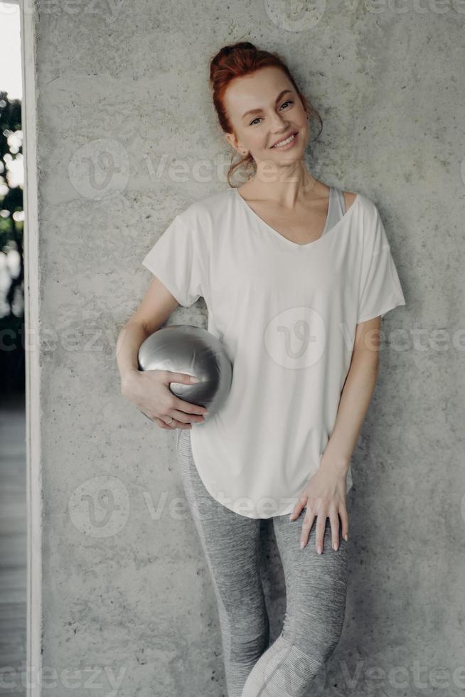 Rothaarige lächelnde Frau, die mit Fitball in der Hand an der Wand steht foto