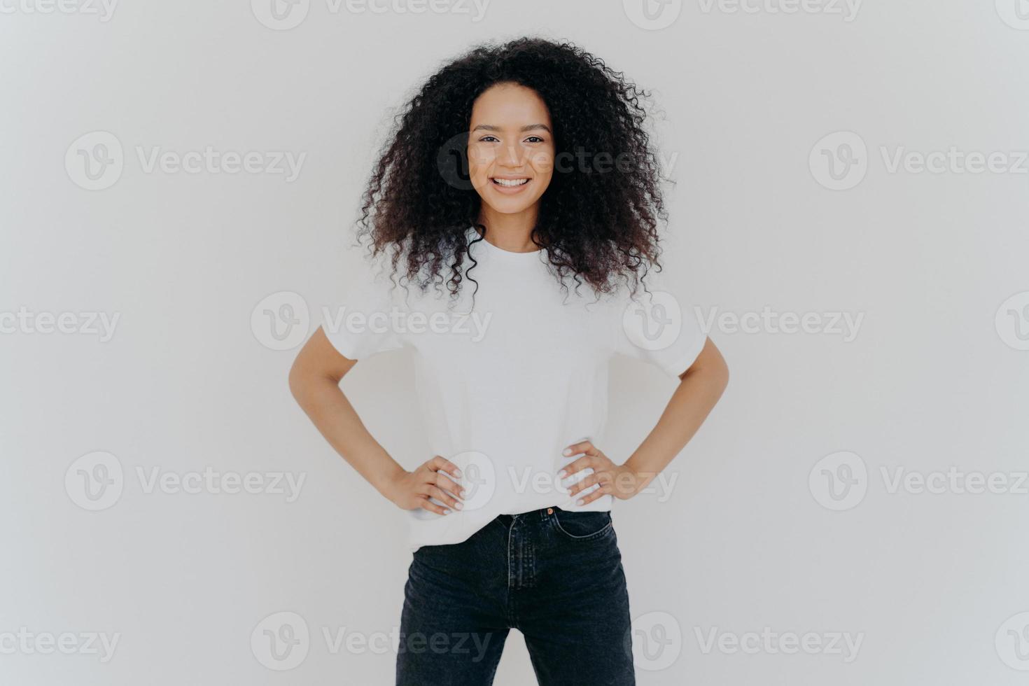 foto der erfreuten lockigen frau hält beide hände auf der taille, lächelt sanft, hat eine schlanke figur, trägt ein weißes t-shirt und schwarze jeans, ist gut gelaunt, steht selbstbewusst vor weißem hintergrund