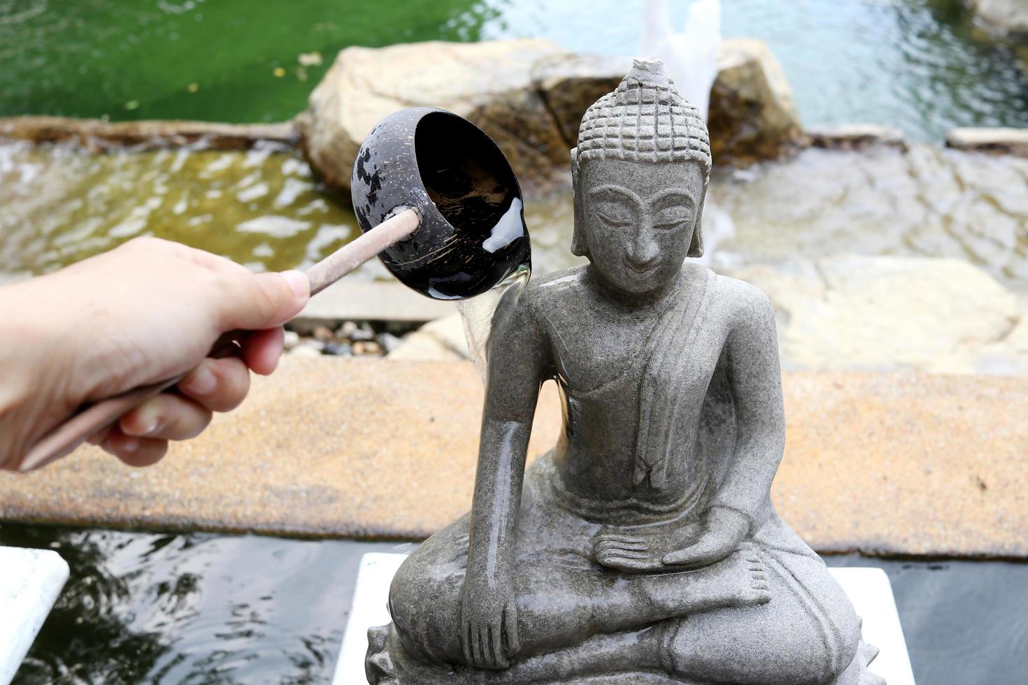 Steingraue Farbe Buddha-Statue neben Teich nass auf der rechten Seite, Wasser fließt aus Kokosnussschalenkelle in der Hand, Thailand. foto