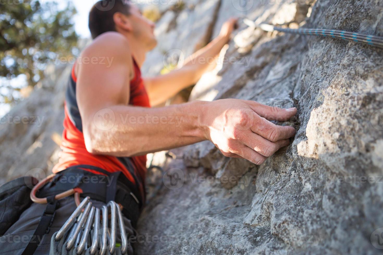 Kletterer im roten T-Shirt klettert auf einen grauen Felsen. Eine starke Hand packte die Führung, selektiver Fokus. Kraft und Ausdauer, Kletterausrüstung Seil, Klettergurt, Chalk, Chalkbag, Karabiner, Hosenträger, Expressschlingen foto