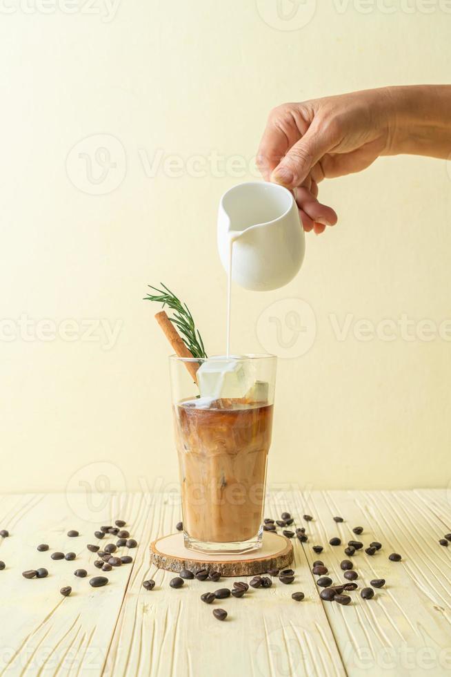 Milch in schwarzes Kaffeeglas mit Eiswürfel, Zimt und Rosmarin gießen foto