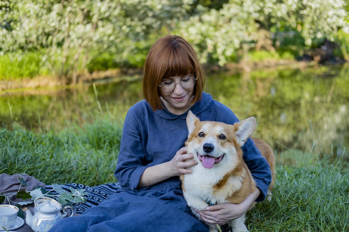 junge frau im retro-kleid mit lustigem corgi-hund auf dem picknick, frau mit süßem hund trinkt englischen tee im park foto