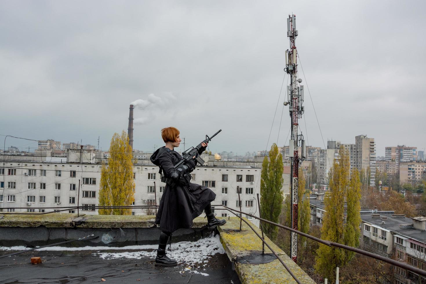 junge frau im modernen schwarzen techwear-stil mit gewehr, das auf dem dach posiert, porträt einer rothaarigen frauen-cyperpunk oder postapokalyptisches konzept foto