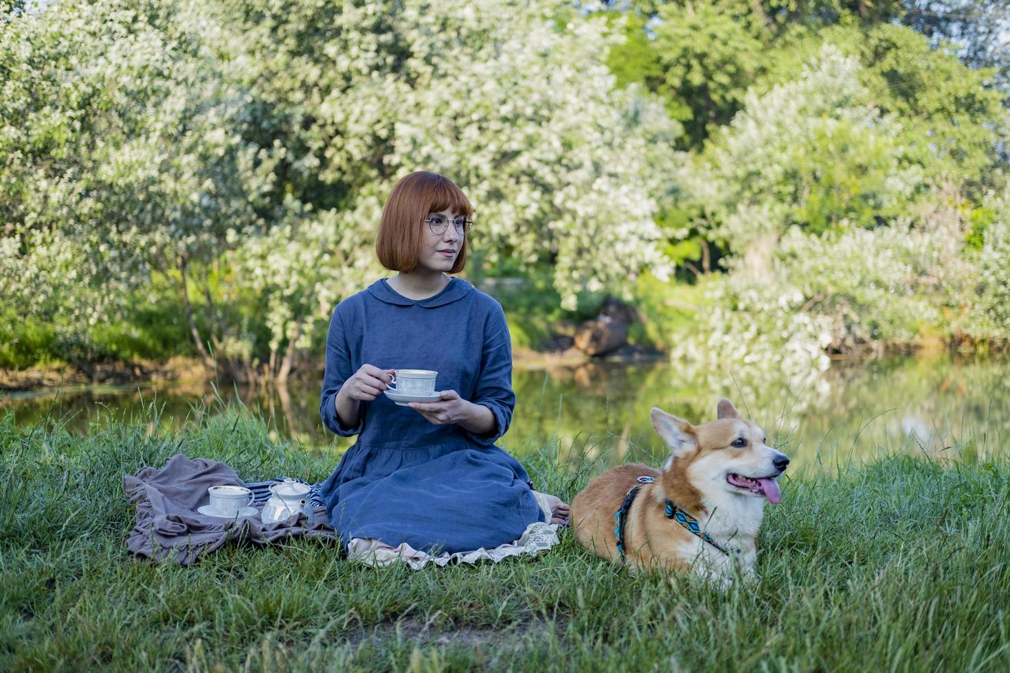 junge frau im retro-kleid mit lustigem corgi-hund auf dem picknick, frau mit süßem hund trinkt englischen tee im park foto