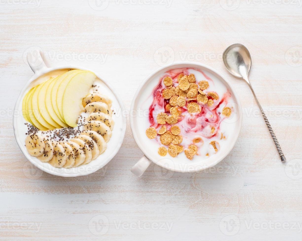 zwei griechischer joghurt mit marmelade, apfel, müsli, chiasamen und banane in weißer schüssel auf weißem holztisch, draufsicht foto