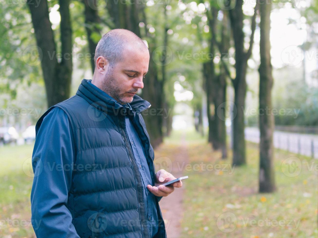 mann hält ein telefon, geht im stadtpark zwischen den bäumen spazieren und schaut auf das smartphone foto
