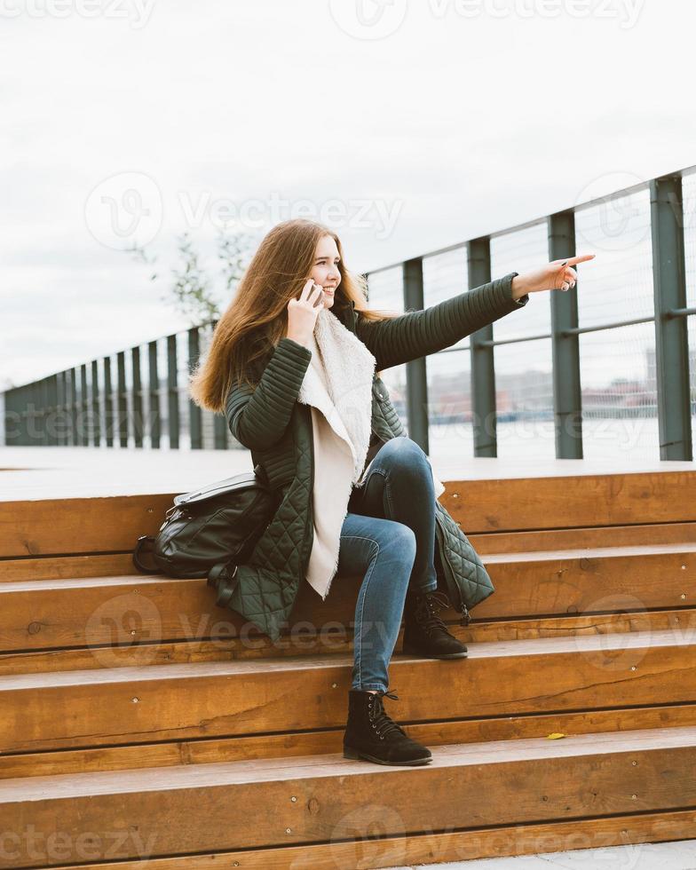 schöne junge frau in warmer kleidung spricht am telefon und zeigt mit der hand. Mädchen sitzt auf Holzstufen am Wasser im Hafen im Herbst, Winter, vertikal foto