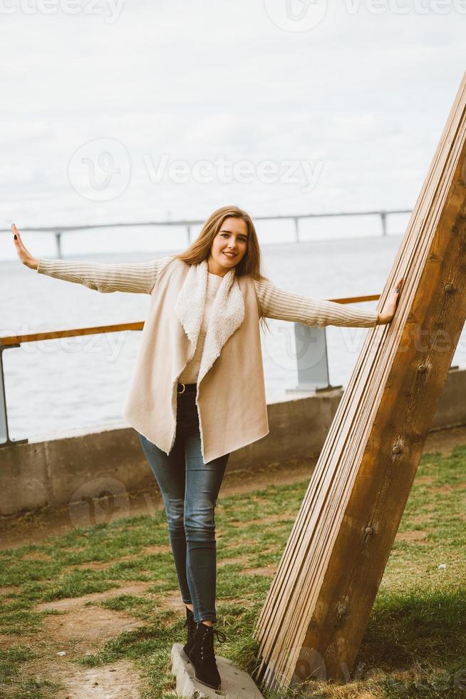 Fröhliches junges Mädchen, das am Wasser am Pier im Hafen steht, das Leben genießt und mit den Armen winkt. Frau mit langen Haaren lächelt und genießt den Moment foto