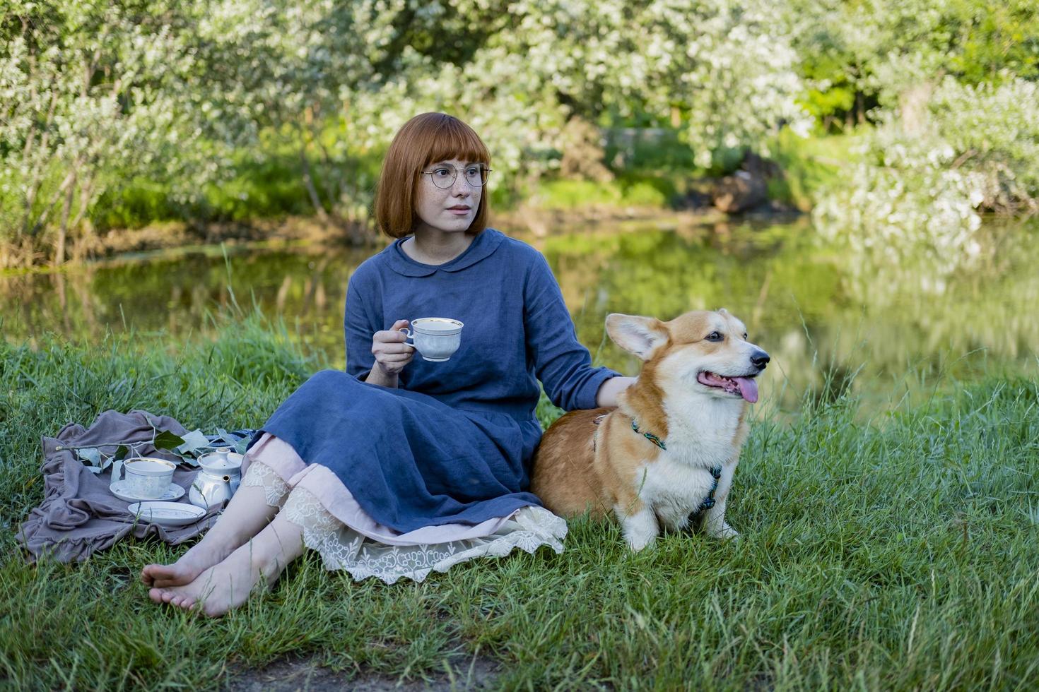 junge frau im retro-kleid mit lustigem corgi-hund auf dem picknick, frau mit süßem hund trinkt englischen tee im park foto