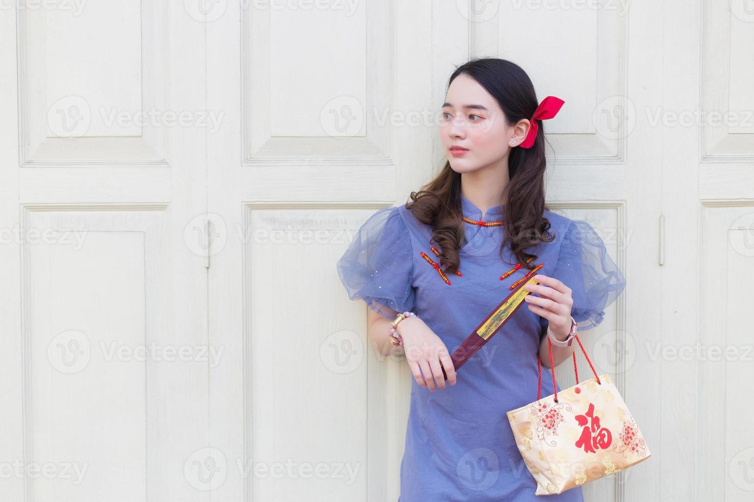 schöne asiatische frau in einem blaugrauen chinesischen kleid, die eine papiertüte mit den worten happy auf chinesisch hält, etwas anschaut und im hintergrund auf einer weißen holztür steht. foto